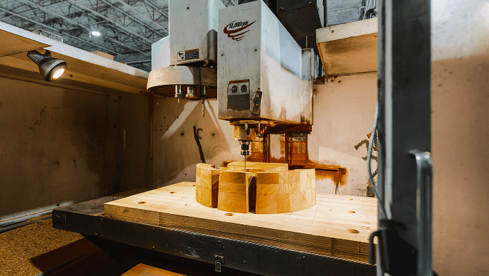 A CNC machine is carving a complex, circular wooden workpiece made of laminated blocks in an industrial setting.