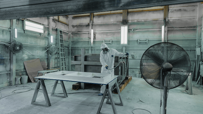  A table in the center of the room with laqer being applied to a metal sheet